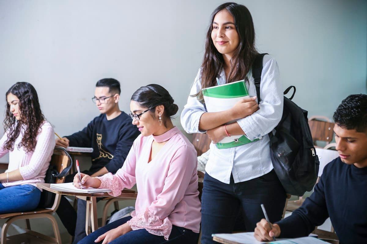 Alunos sorrindo em sala de aula, com uma estudante em pé segurando um caderno